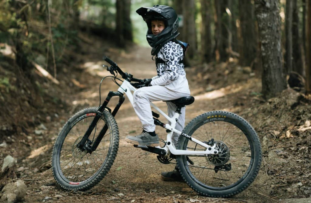 Copil pe bicicleta mountain bike full-suspension pe poteca din padure, cu casca si echipament MTB